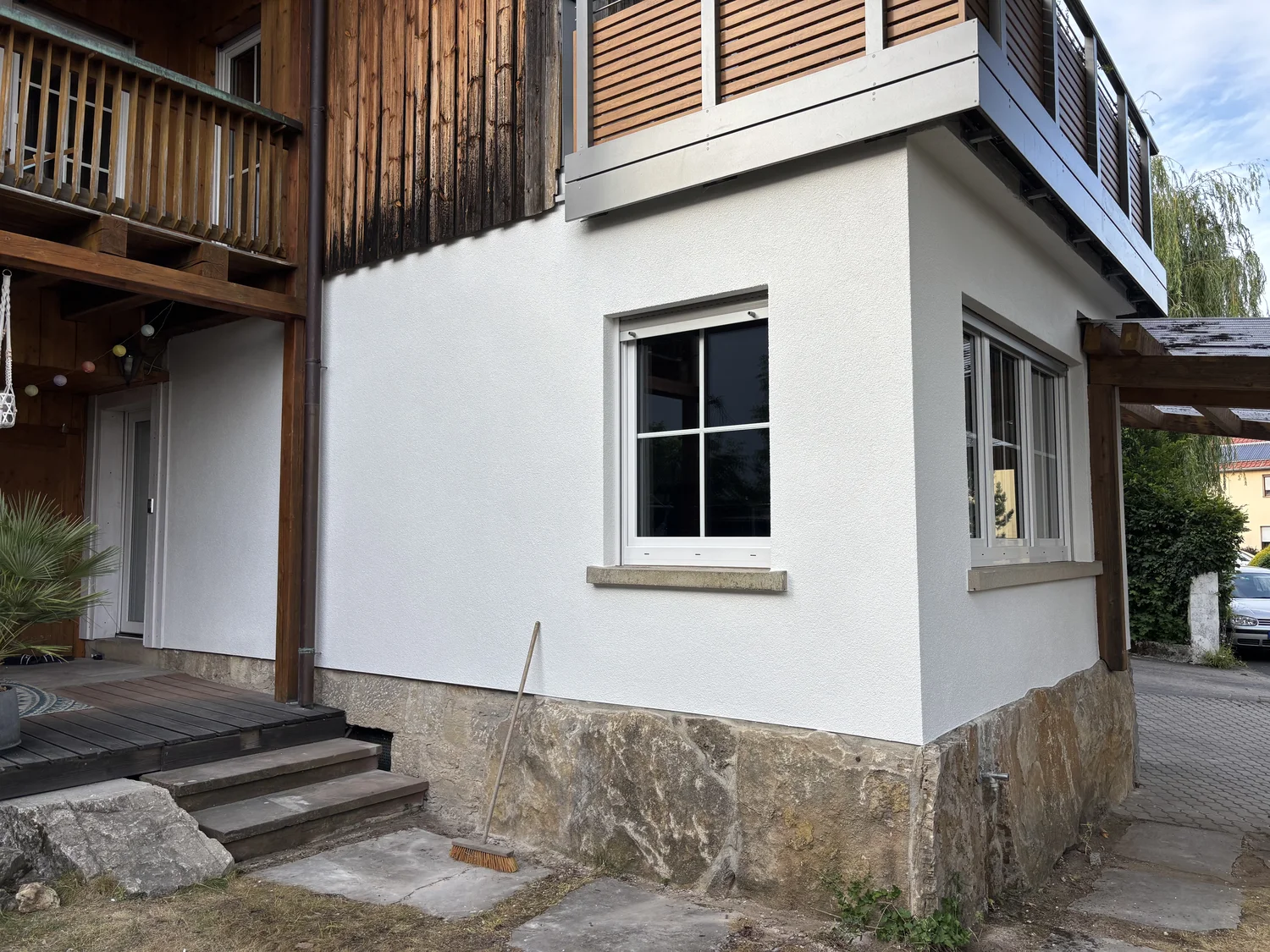 M. Ulusoy & Söhne GbR – Malermeisterbetrieb – Ecke eines modernen Hauses mit weiß verputzten Wänden, Steinfundament, Holzbalkon und zwei weiß gerahmten Fenstern