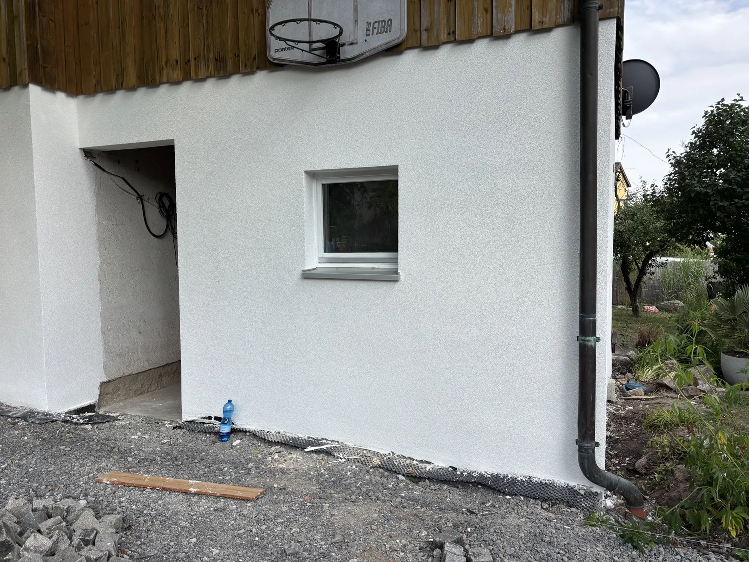 M. Ulusoy & Söhne GbR – Malermeisterbetrieb – Teil einer weißen Hausaußenwand mit einem kleinen Fenster und einem darüber hängenden Basketballkorb, eine blaue Wasserflasche auf dem Boden neben Kies und Baumaterialien
