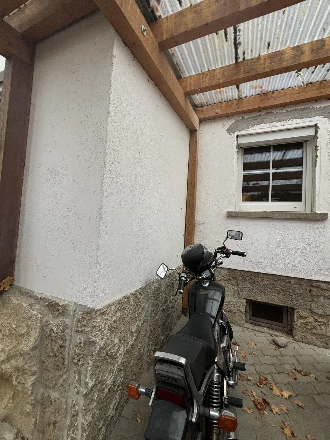 M. Ulusoy & Söhne GbR - Malermeisterbetrieb - Schwarzes Motorrad parkt unter einer Holzkonstruktion neben einer weißen Steinfassade mit geschlossenem Fenster