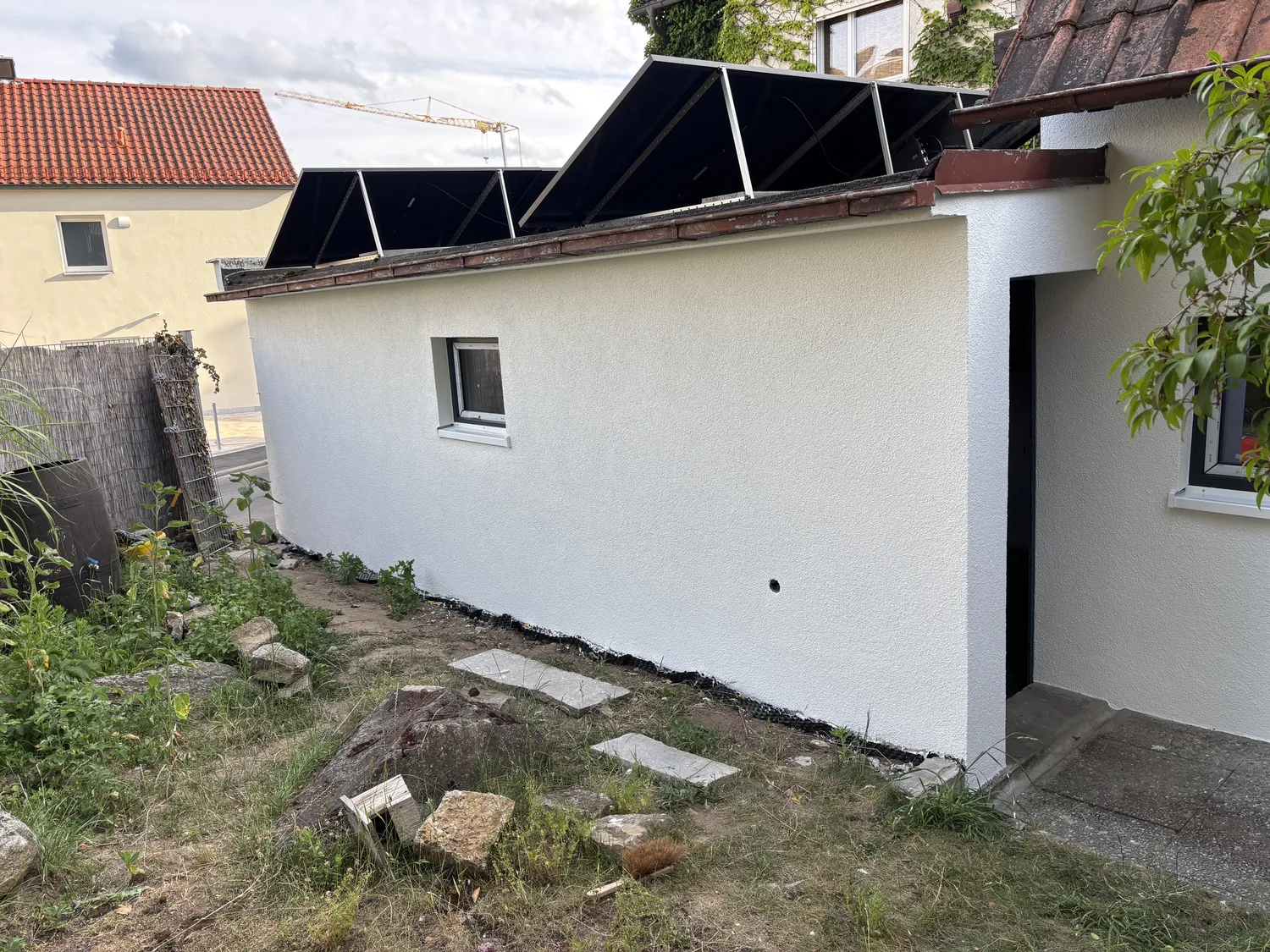 M. Ulusoy & Söhne GbR – Malermeisterbetrieb – Weißes Gebäude mit auf dem Dach montierten Solarpaneelen und einem kleinen Fenster an der Seite, umgeben von Gras, Steinen und Gartenpflanzen