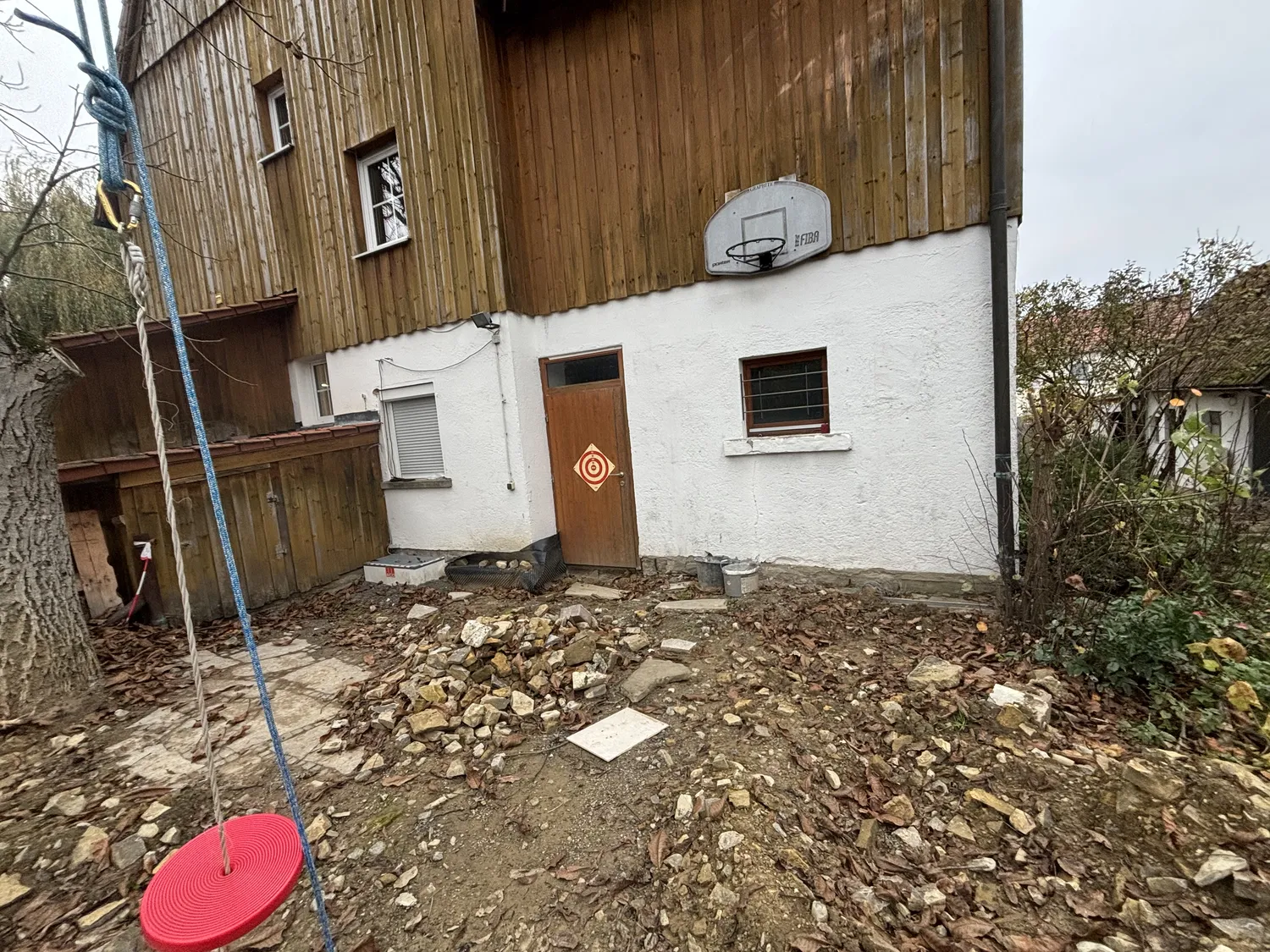 M. Ulusoy & Söhne GbR – Malermeisterbetrieb – Hinterhofbereich mit einer Hausfassade aus Holz und weißem Stuck, einem Basketballkorb über einer Holztür, einer roten Schaukelscheibe, die an einem Baum hängt, und Haufen von Steinen und Erde auf dem Boden