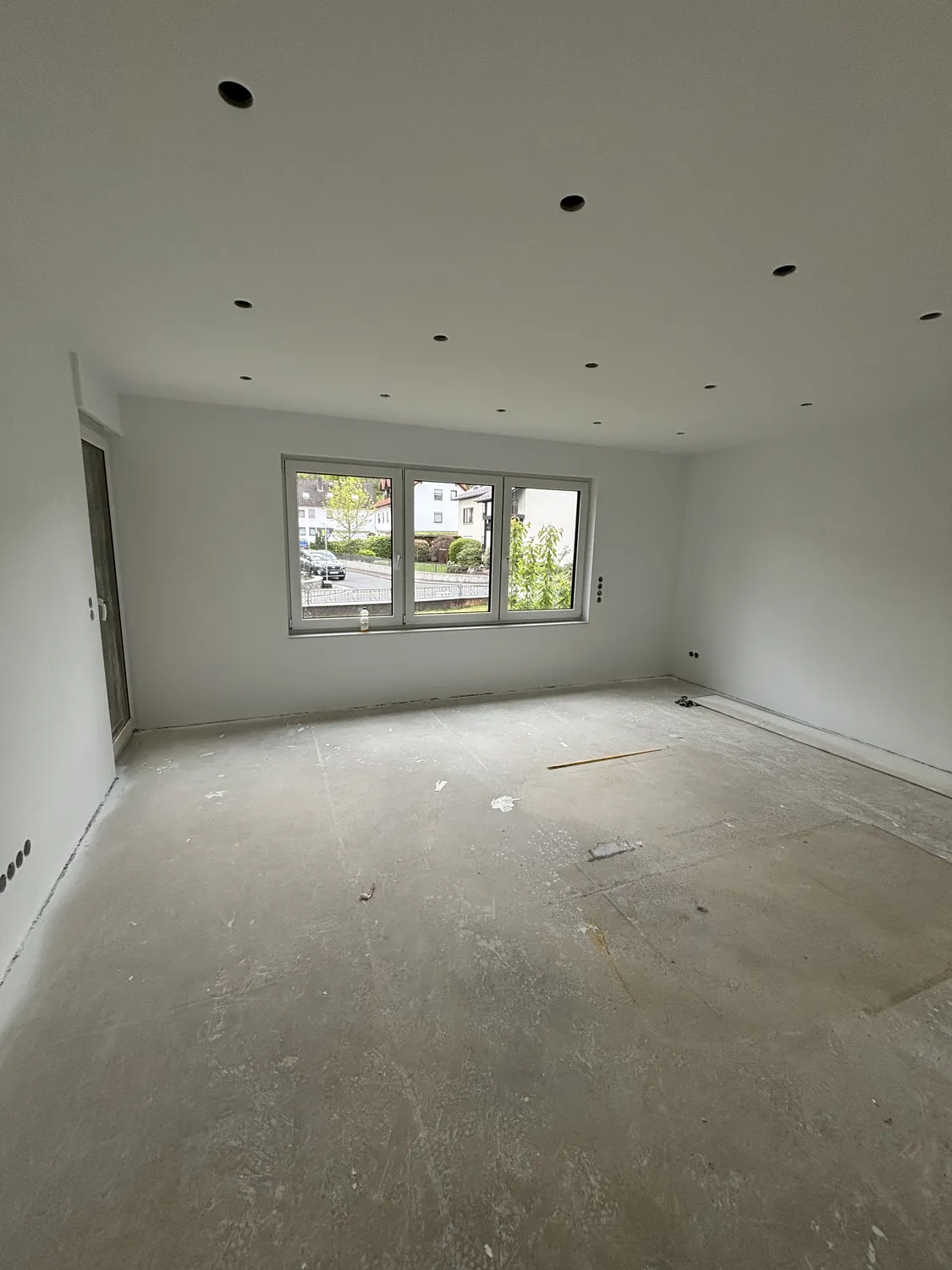 Empty room under construction with bare concrete floor, white walls, ceiling lights, and a three-panel window showing an outdoor street view.