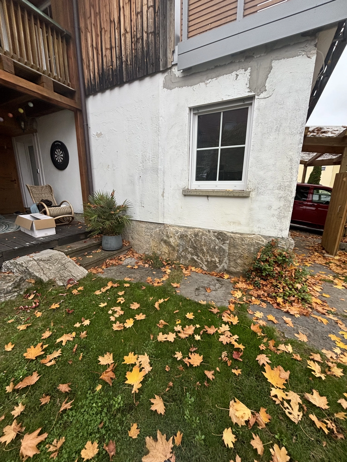 M. Ulusoy & Söhne GbR – Malermeisterbetrieb – Herbstlaub verstreut auf grünem Gras an der Ecke eines weißen Hauses mit Fenster und einer kleinen Topfpflanze in der Nähe