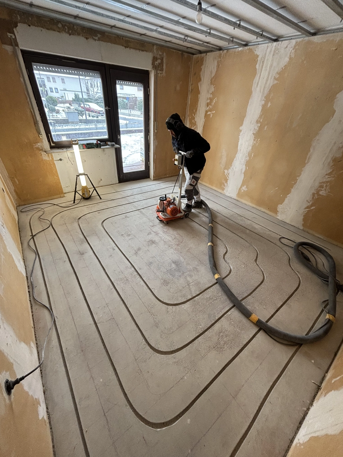 M. Ulusoy & Söhne GbR – Malermeisterbetrieb – Arbeiter bedient eine Bodenfräse, um Rillen in einen Betonboden in einem Rohbauraum mit Fußbodenheizungsanlage zu schneiden.
