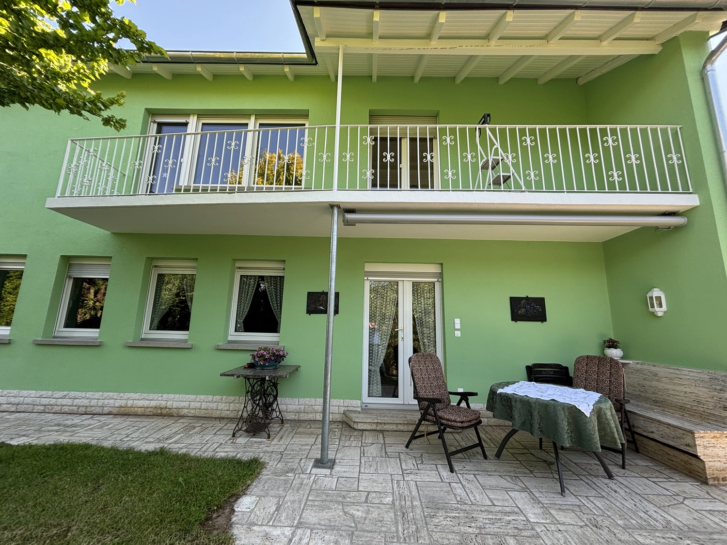 M. Ulusoy & Söhne GbR – Malermeisterbetrieb – Grünes zweistöckiges Haus mit weißem Balkongeländer, Terrassentisch mit Stühlen, Topfpflanzen und Steinboden